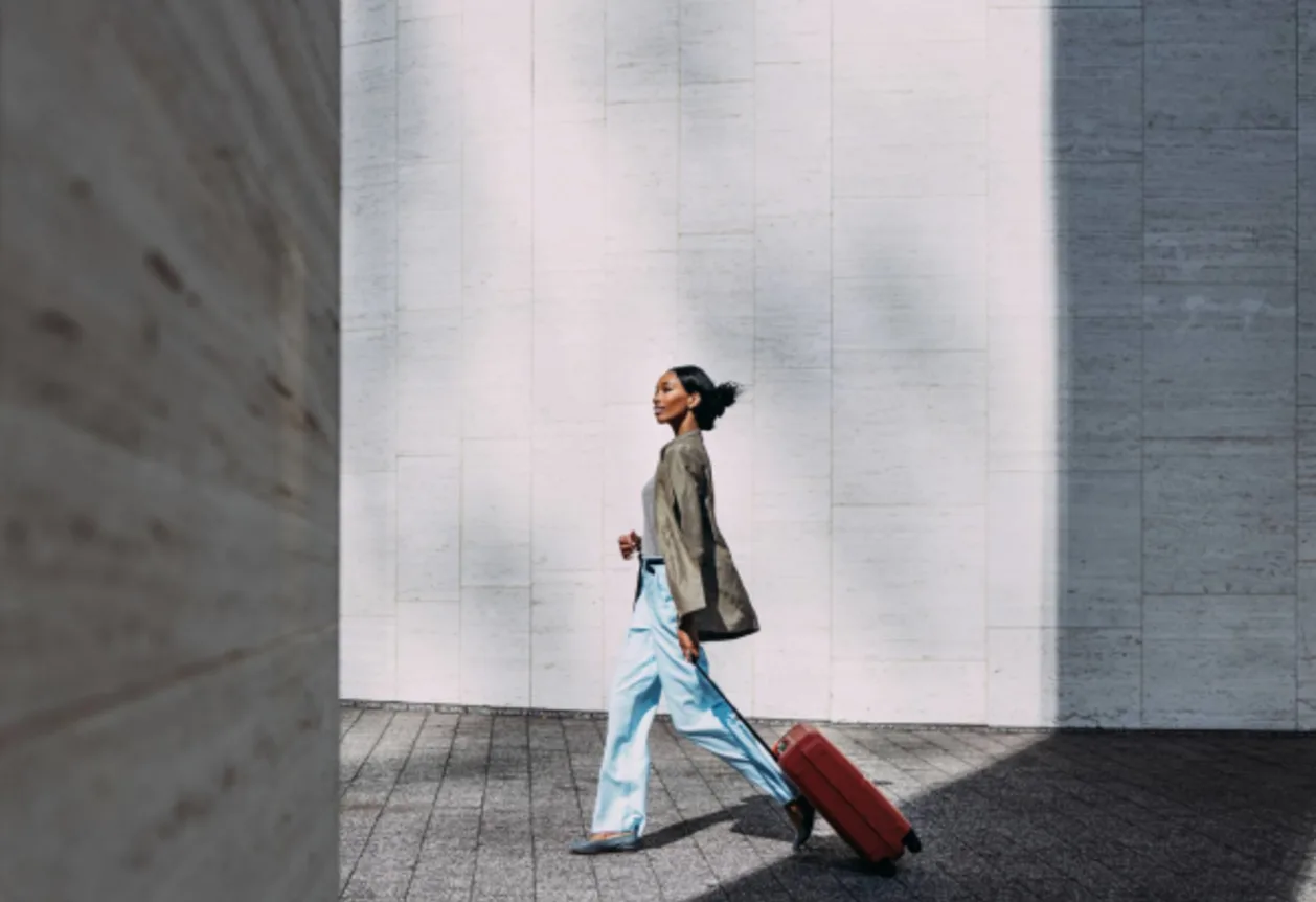 Confident black woman walking with red suitcase against a modern urban wall